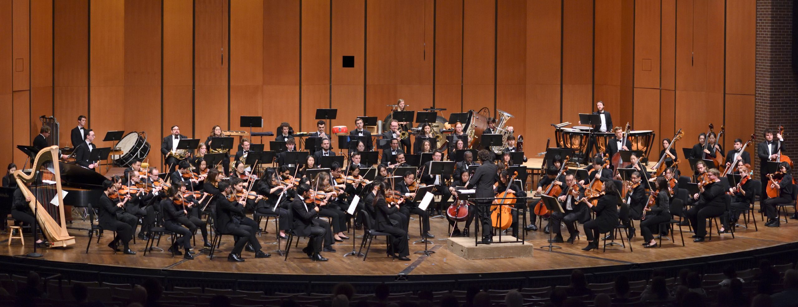 Wide shot of MSU Orchestra playing on stage in front of music stands. There are chairs and a variety of instruments set up around the stage. There is a conductor in the middle.