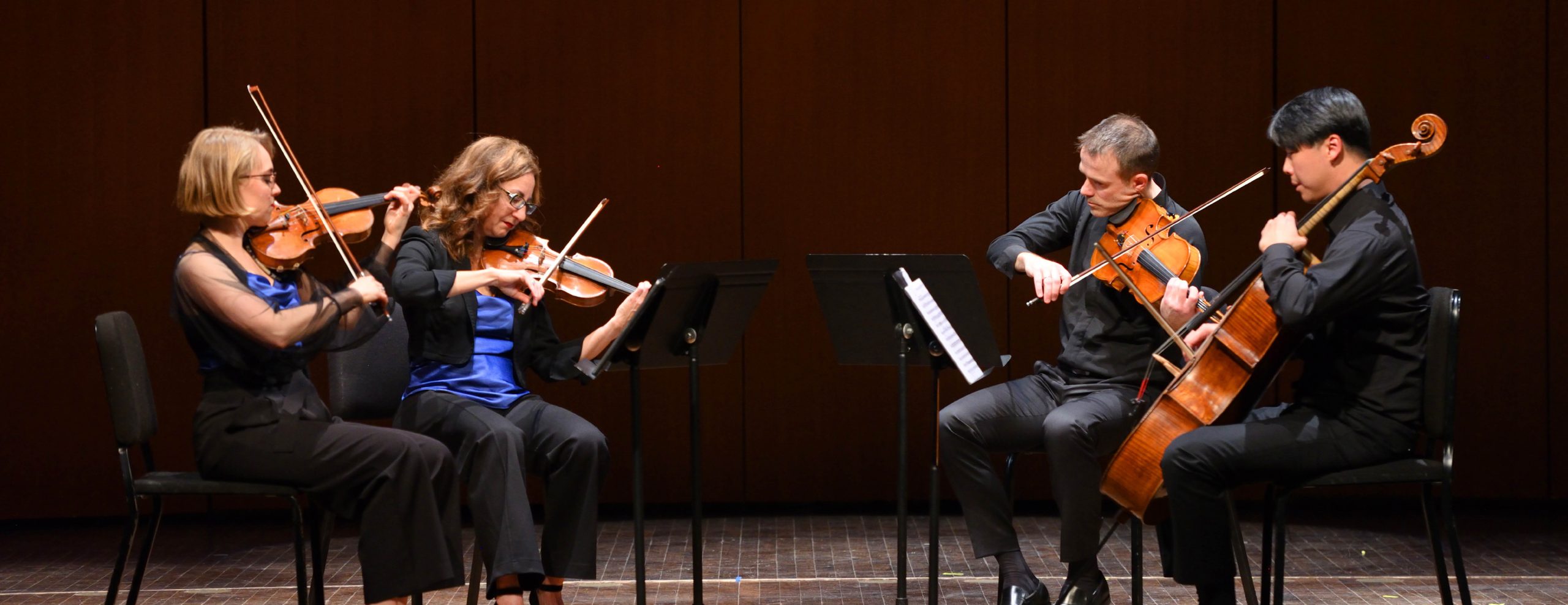 Image shows a string quartet playing their instruments on stage. They are sitting in chairs with music stands. Their bows are up.