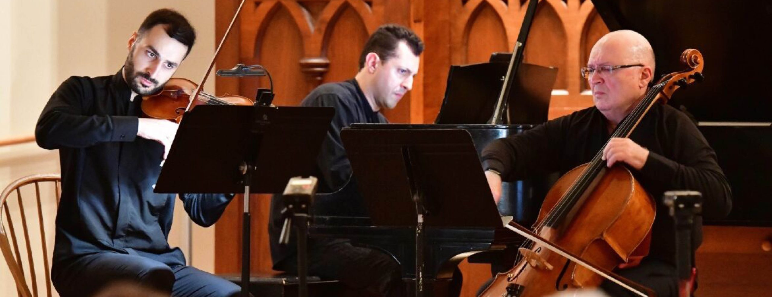 Image shows three musicians playing together on stage. The instruments being played are the cello, violin and piano.