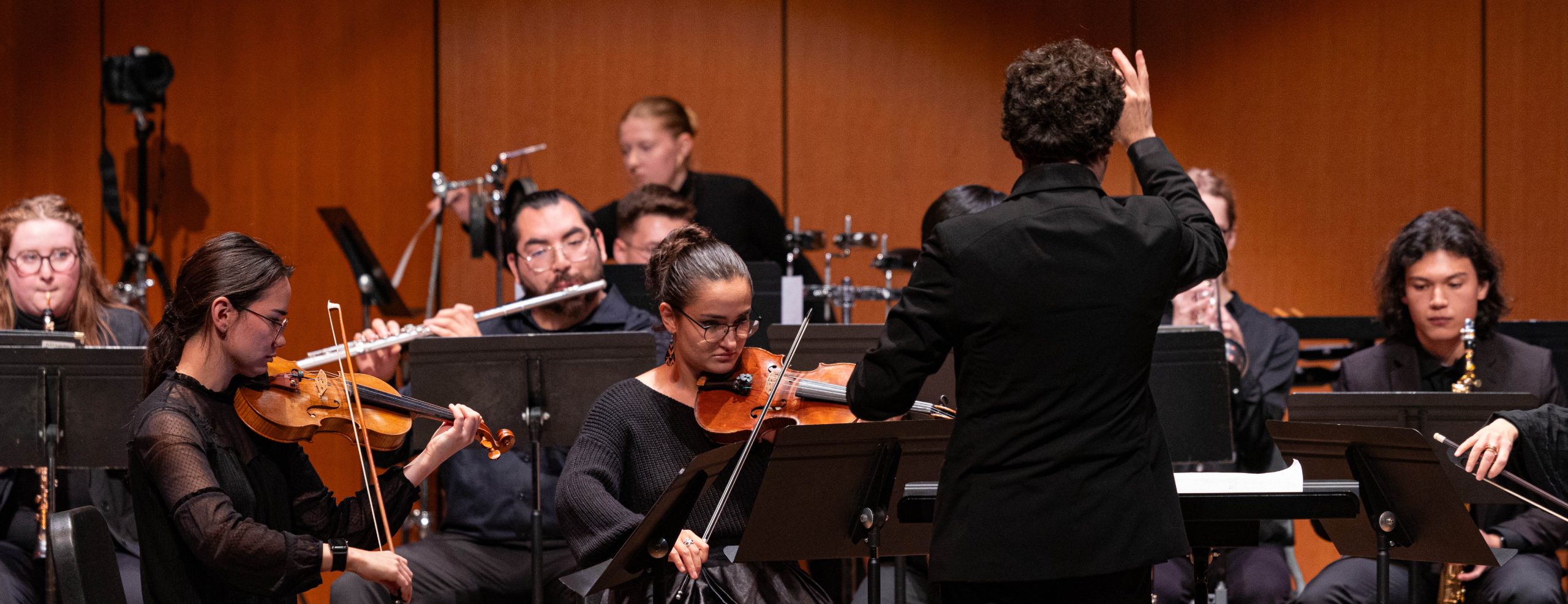 A variety of student musicians are playing their instruments on stage in front of a conductor.