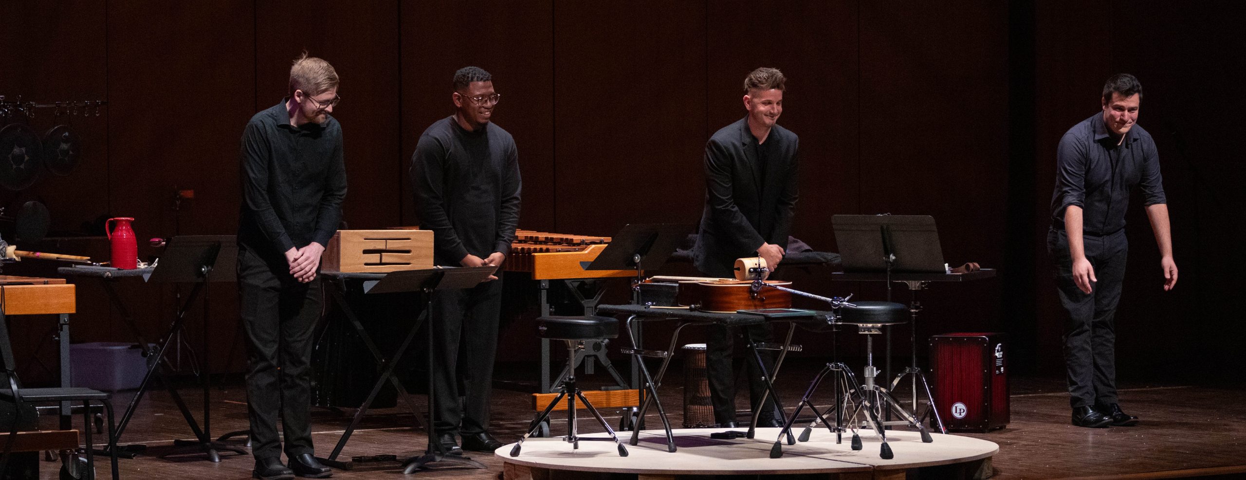 Four percussion musicians taking their bows on stage. Variety of percussion instruments around the stage.