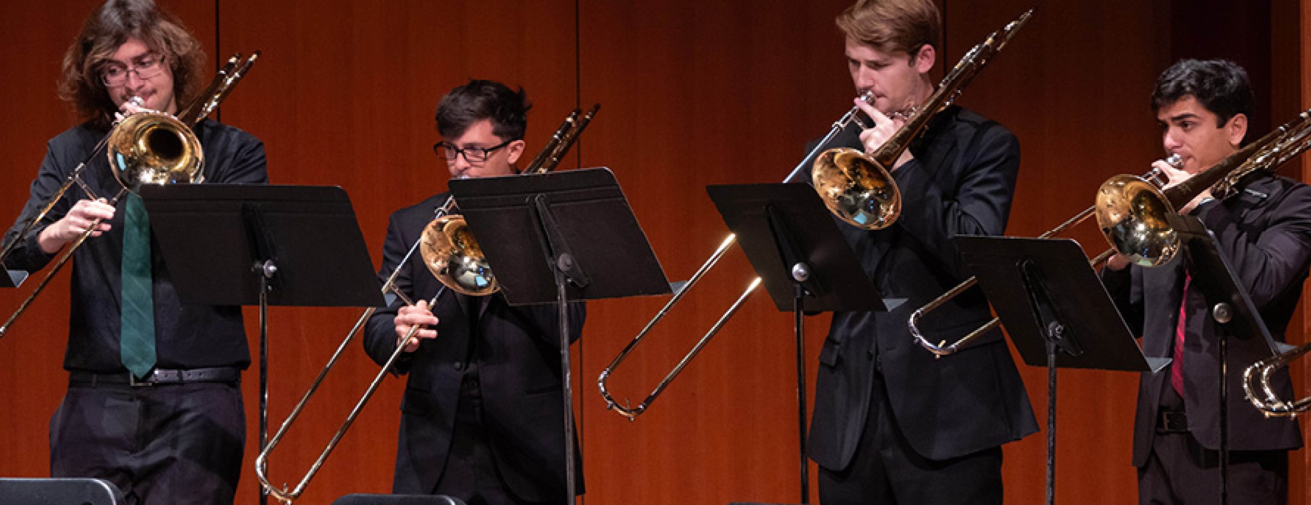 Four individuals playing the trombone, dressed in formal attire. They each have a music stand in front of them.