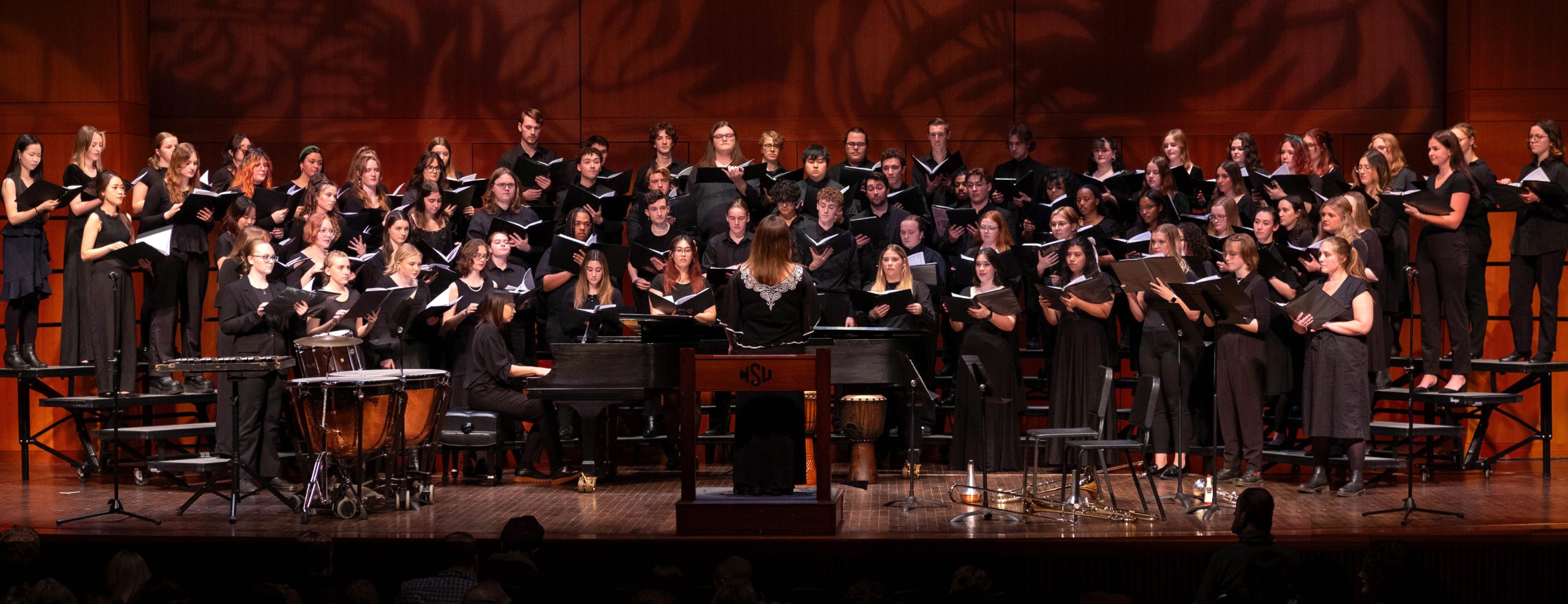 A wide image of a choir dressed in all black performing on stage in front of a conductor. They are standing on risers and there is stage equipment and instruments in front of them.