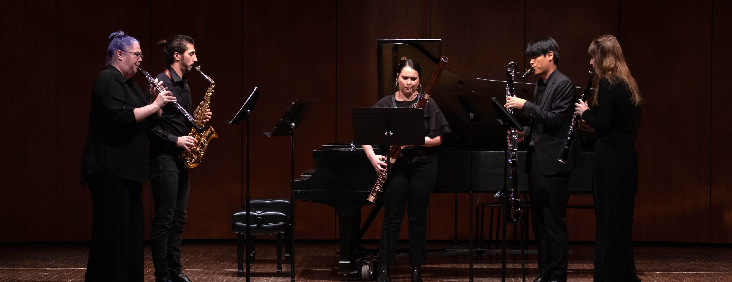 Five woodwind musicians playing their instruments on stage. There is a grand piano in the background behind them. There are music stands in front of each musician.