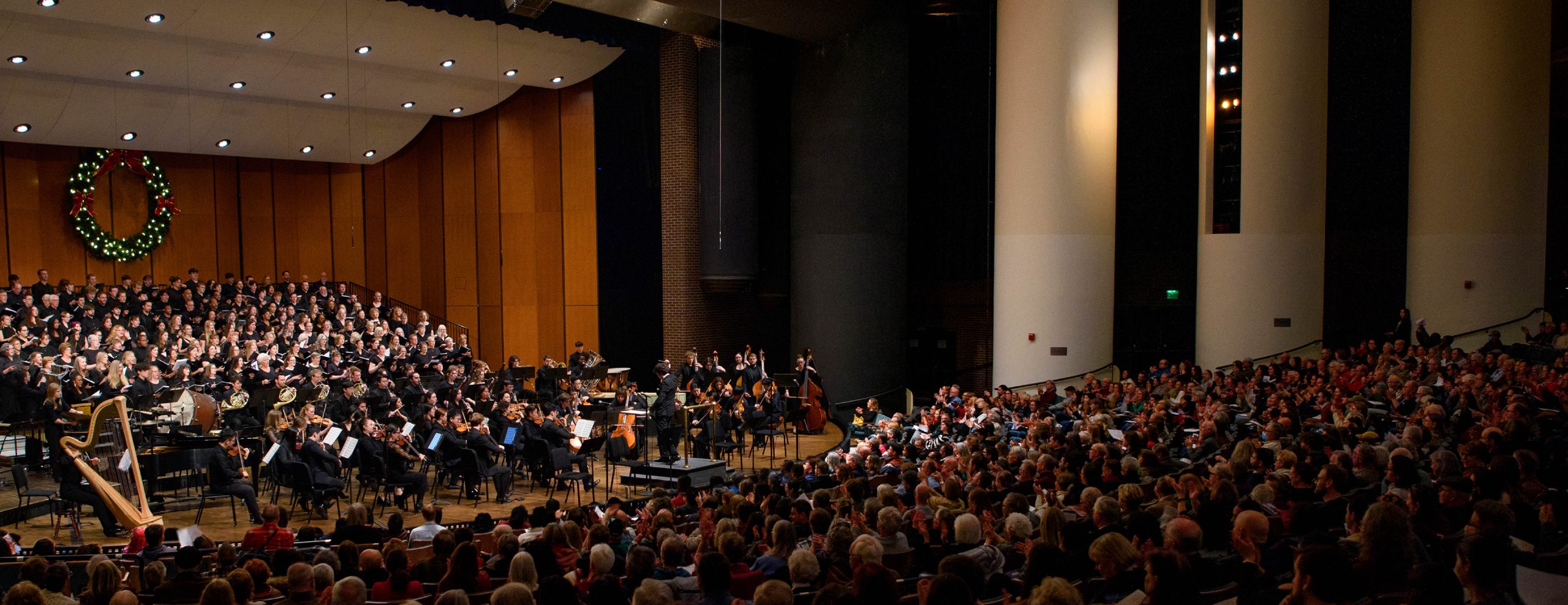A wide, zoomed out image. On the left, MSU choirs and orchestra come together to perform on stage. On the right, the audience is watching the performance.