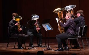 Two tuba students and two euphonium students play their instruments on stage in front of music stands.
