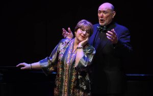 2 vocalists performing on stage holding binders with music. Behind them is a grand piano with a pianist playing it.
