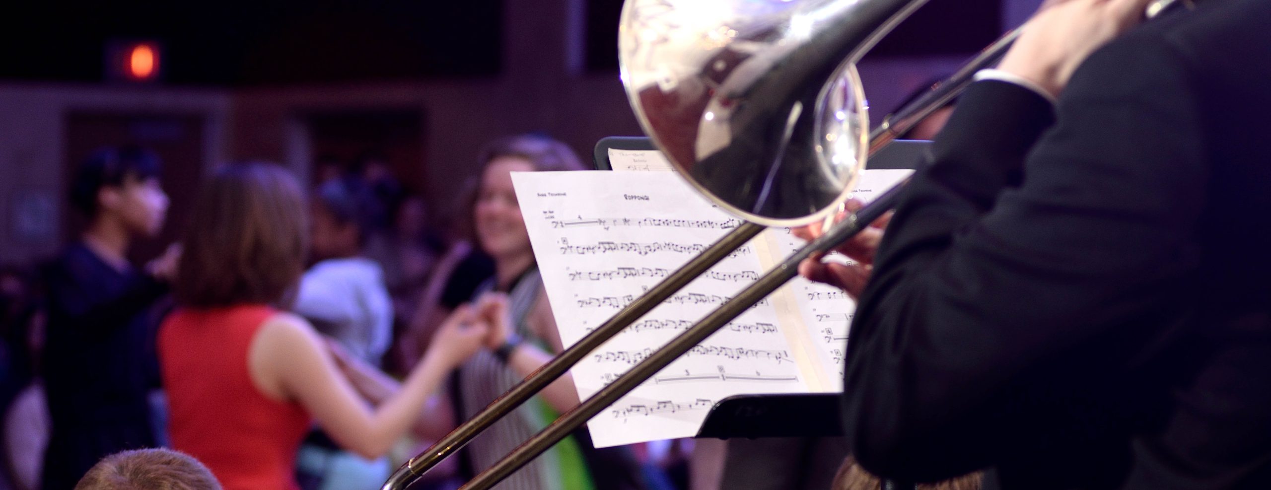 Close up of MSU Jazz student playing the trombone in front of music stand with sheet music on it. Background shows people doing swing dance.