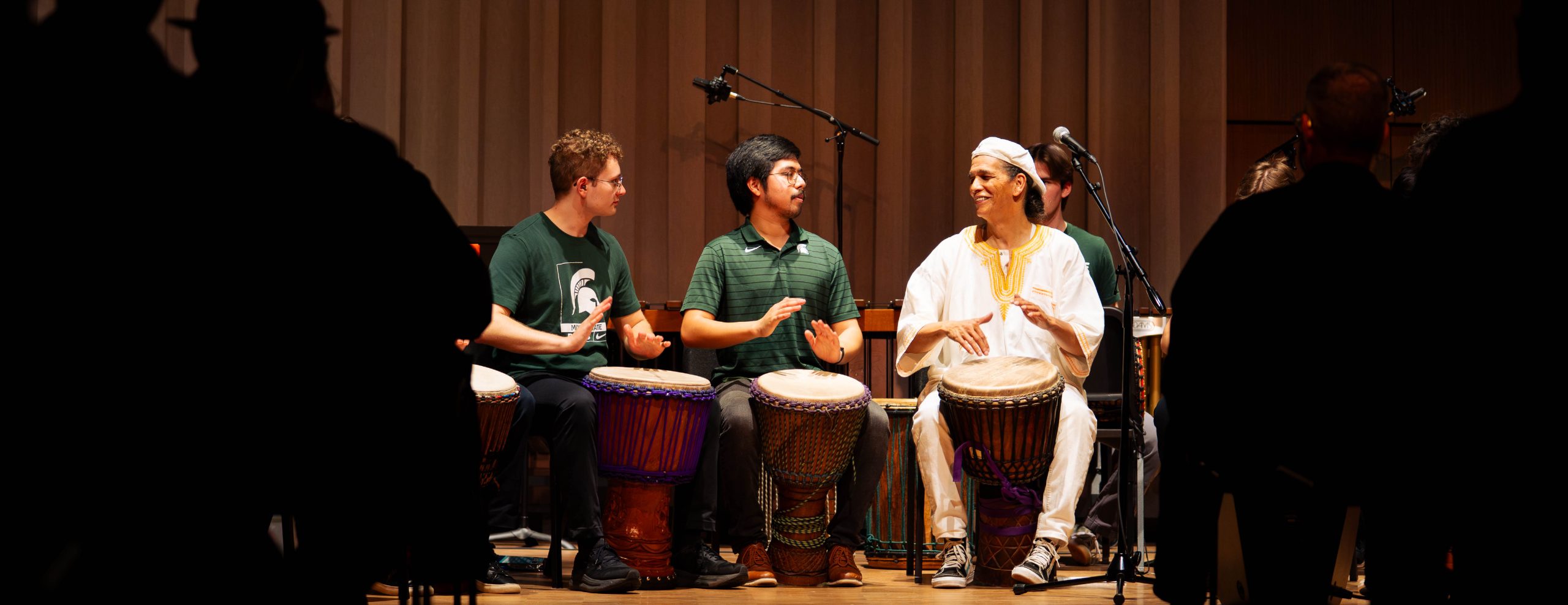 African Diaspora Percussion Ensemble performing on stage. The image is framed by shadows of an audience. In the center, there are three individuals playing drums.