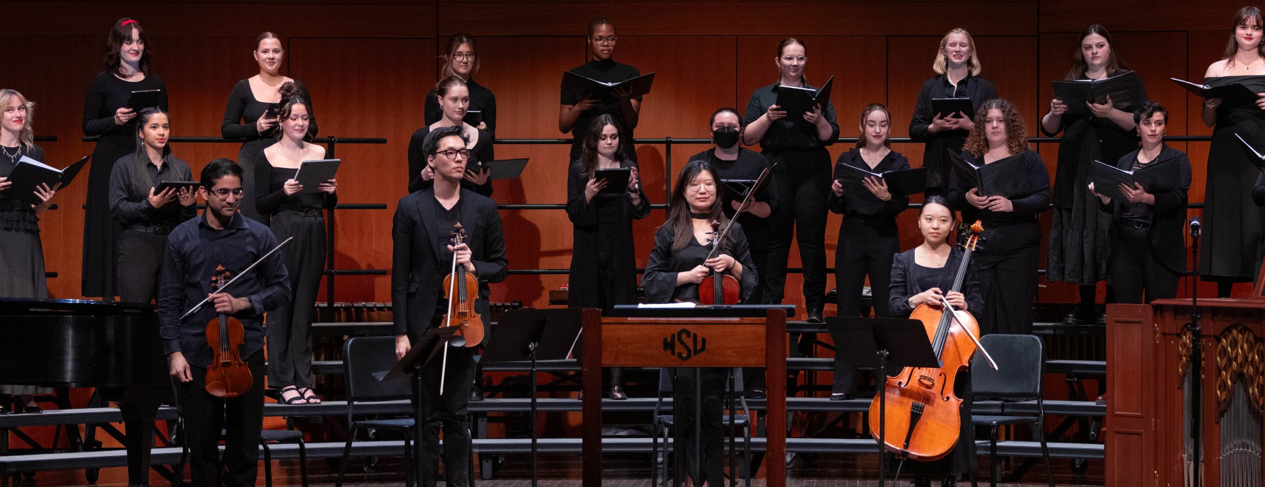 A choir dressed in all black performing on stage. In front of them, there are four string players standing with their instruments for applause.