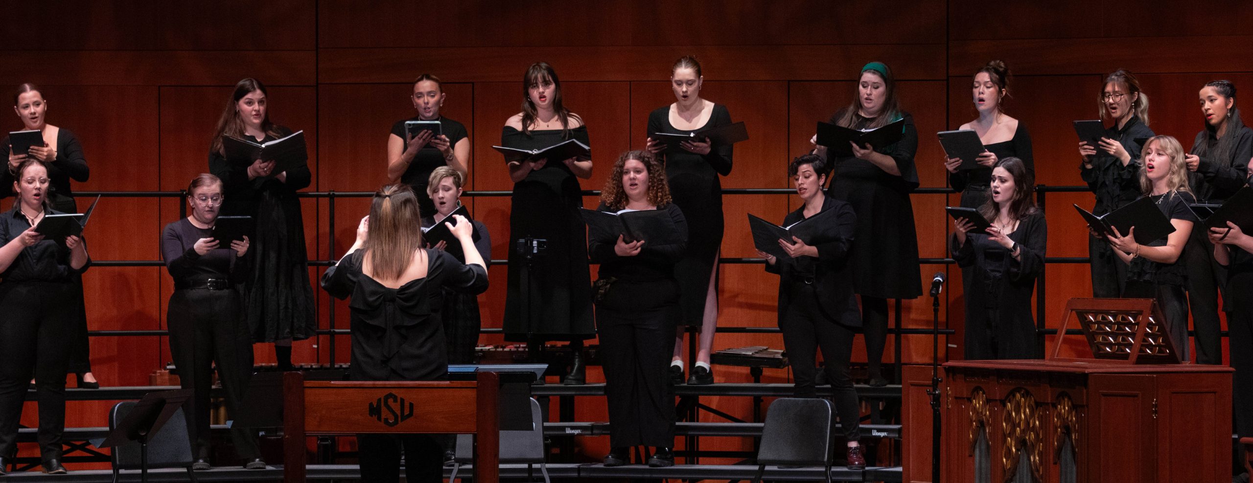 MSU Choir Mosaic performing on stage dressed in all black, in front of a conductor.