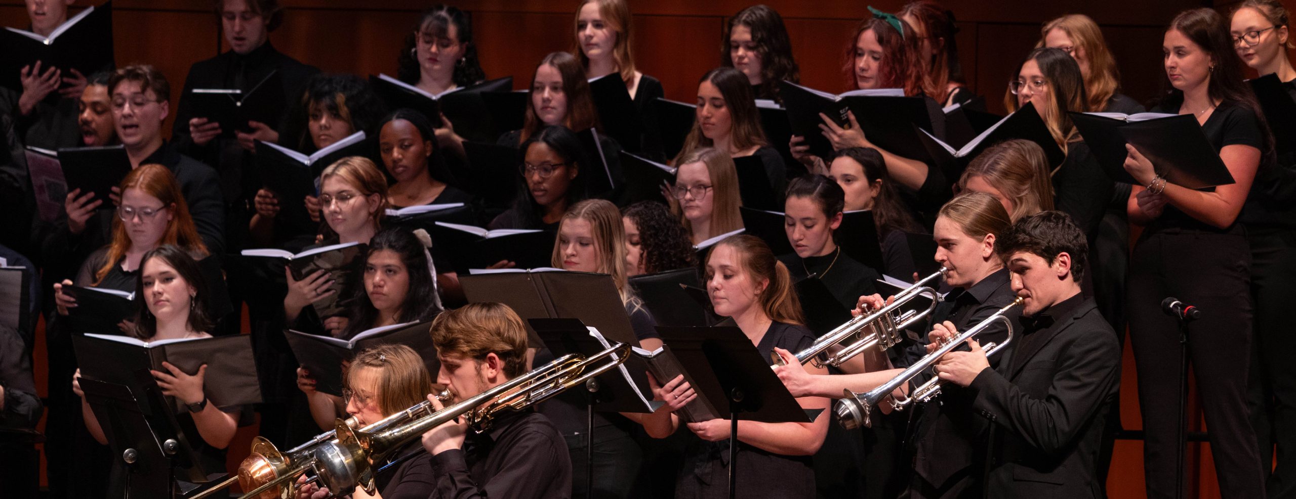 Choir and trumpet players on stage performing together. The trumpet players are in the foreground and the choir is in the background.