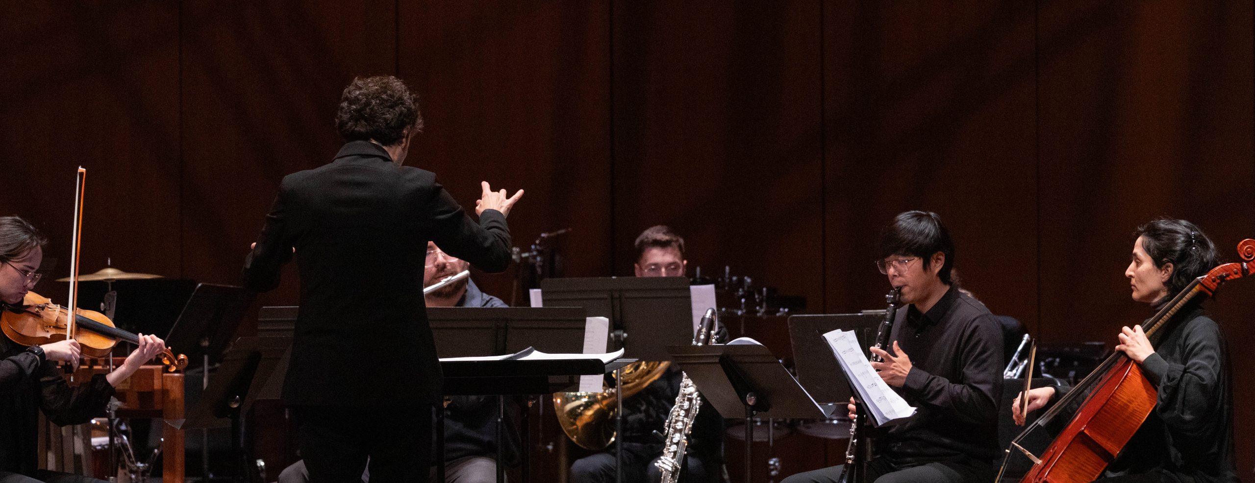 Variety of student musicians including cello, oboe, violin and flute are playing instruments on stage in front of a conductor.