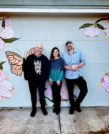 three people stand with their backs against a wall painted with butterflies