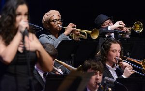 Closeup of horn section of jazz orchestra performing, in foreground to the left, jazz vocalist is singing.