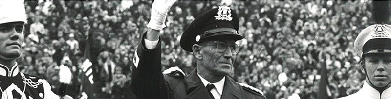 black and white image of person in military style marching band uniform waving to a crowd with two band members to each side