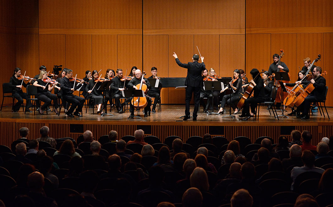 medium sized orchestra of string musicians gathered on stage for performance and led by a conductor in Fairchild Theatre. Full audience in the foreground