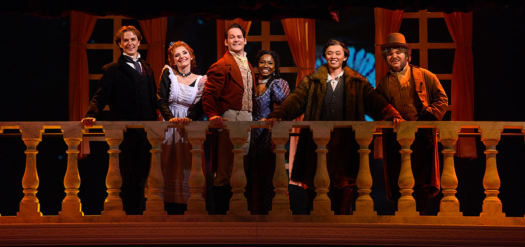 Six opera cast members standing at the top of a decorated balcony platform, smiles are their faces, formerly dressed in costumes.