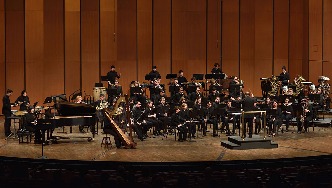 Large ensemble onstage, wind-band instrumentalists performing a variety of instruments, from percussion to brass, to woodwind. Conductor in foreground leading the group.