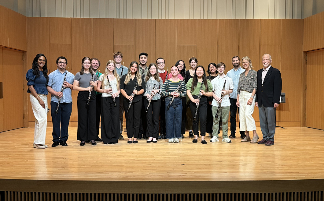 a group of 19 people side to side across a stage, most of them holding oboes