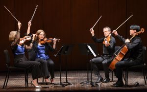 Image shows a string quartet playing their instruments on stage. They are sitting in chairs with music stands. Their bows are up.