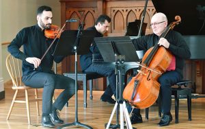 Image shows three musicians playing together on stage. The instruments being played are the cello, violin and piano.